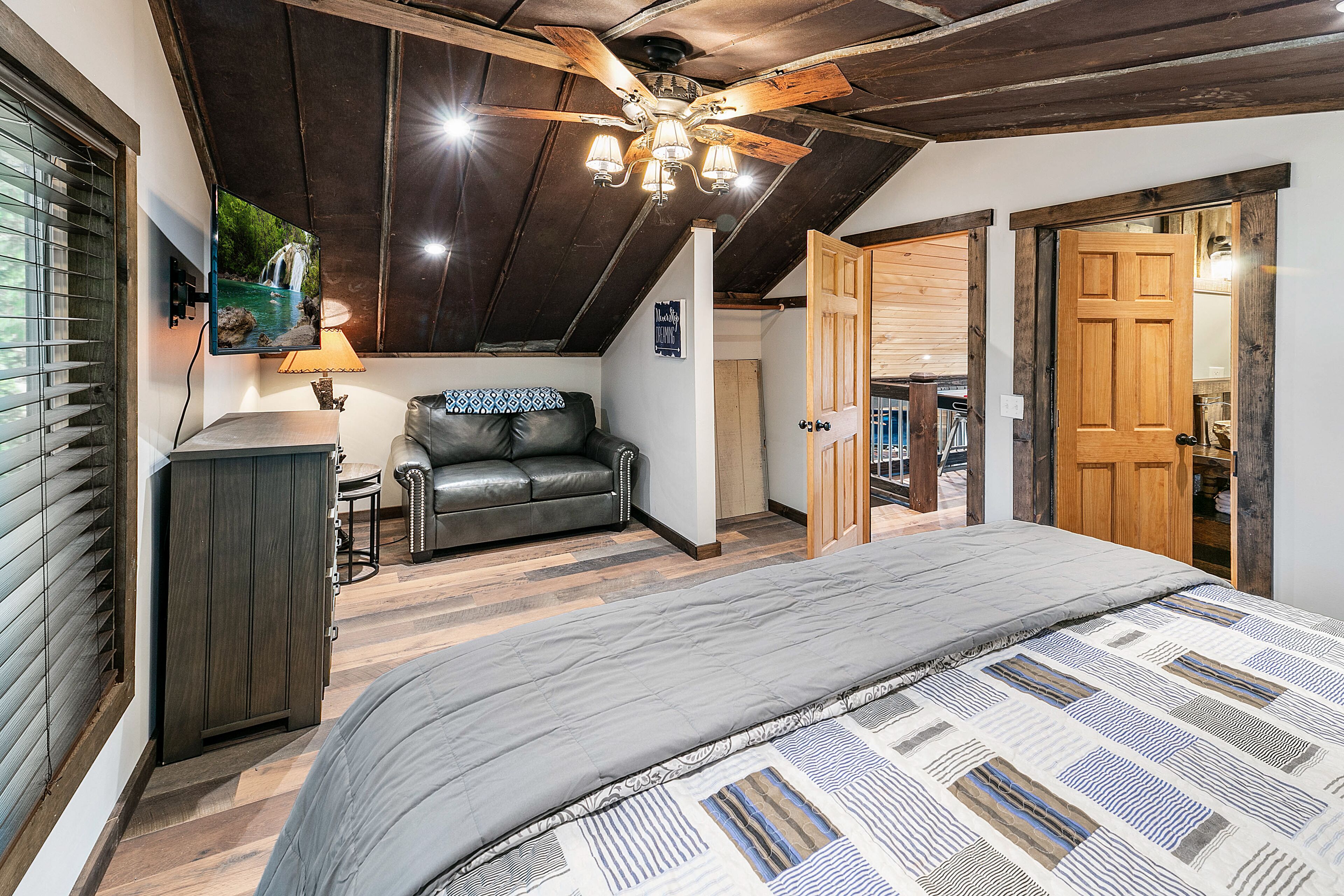 A bedroom with a large bed, a ceiling fan, a TV mounted on the wall, and a wooden door leading to another room.