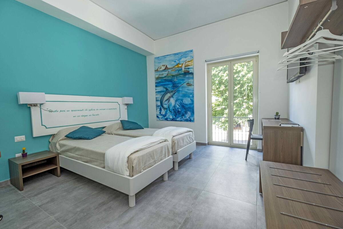 A bedroom with two beds, a desk, a chair, a window, and a painting on the wall.
