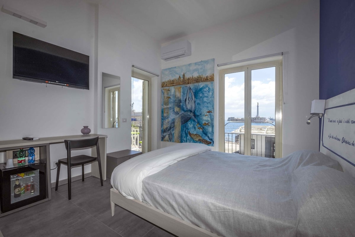 A bedroom with a bed, a desk, a chair, a TV, and a painting.