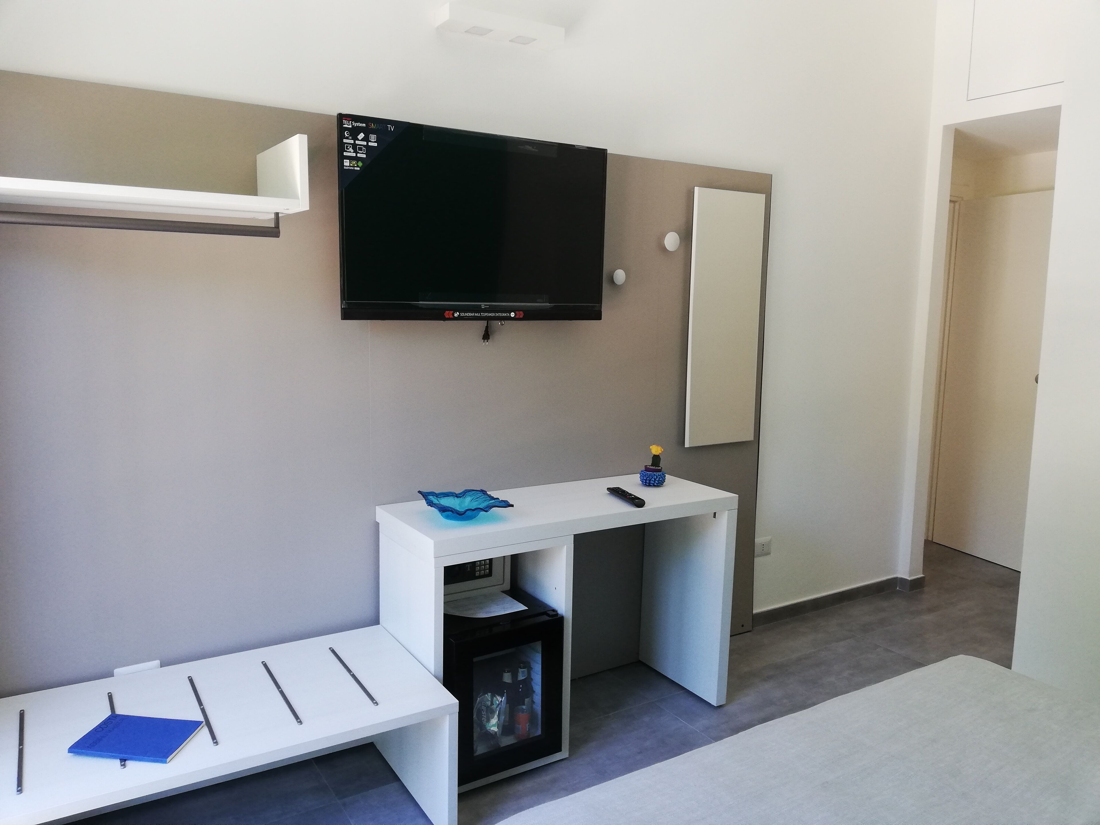 Una habitación moderna con televisor de pantalla plana en la pared, un escritorio blanco con control remoto, un pequeño florero con una flor y un microondas.