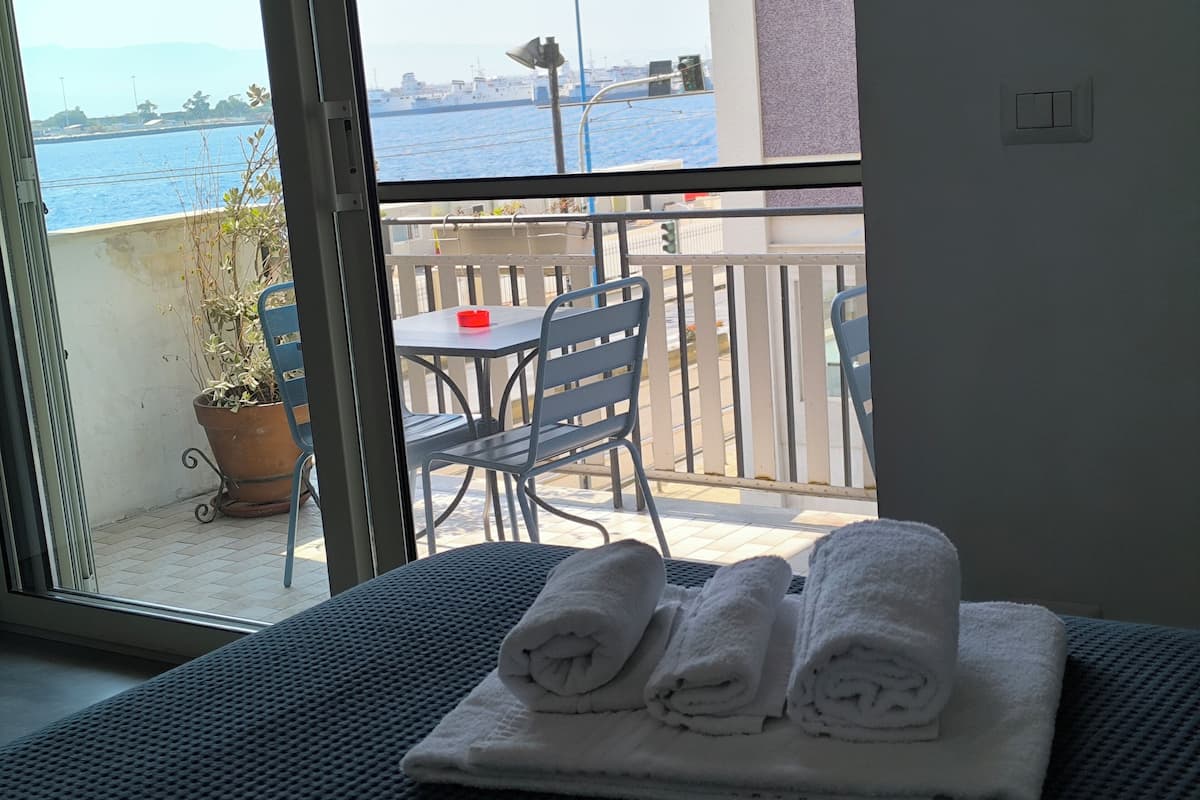 A balcony with a table and chairs, a potted plant, and a view of the sea and ships.