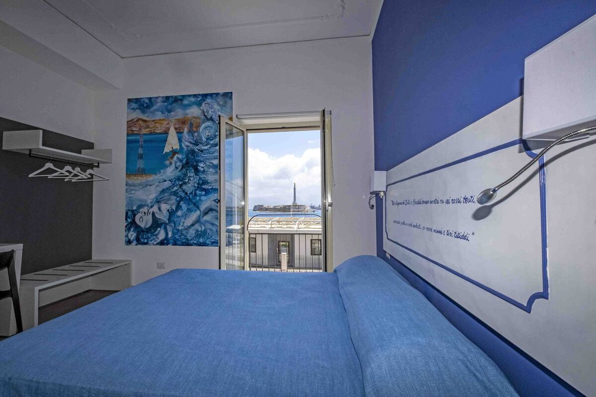 A bedroom with a bed, a painting of a seascape, a window with a view of a building, and a wall-mounted towel rack.