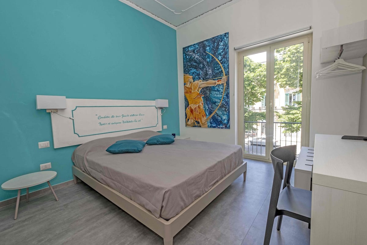 A modern bedroom with a bed, a desk, and a chair. There is a window with a view of trees and buildings.
