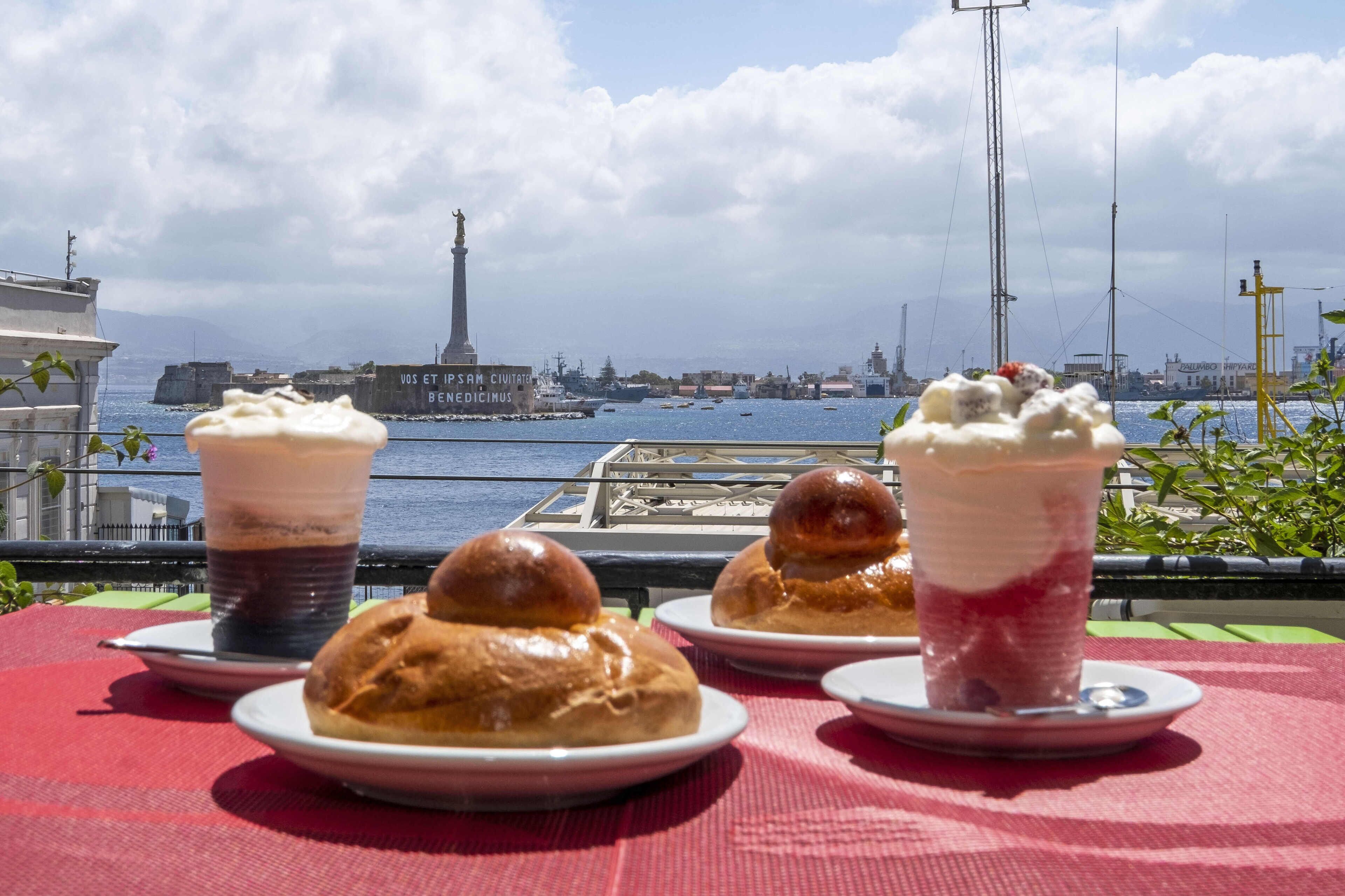 Un tavolo con dolci e bevande, affacciato su un porto con navi e un monumento.