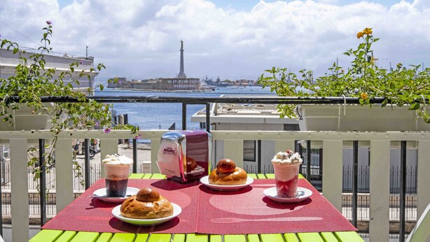 A balcony table with food and drinks, overlooking a body of water and a tower in the distance.