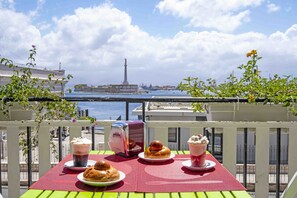 A balcony table with food and drinks, overlooking a body of water and a tower in the distance. - Miti dello Stretto B&B (Messina)