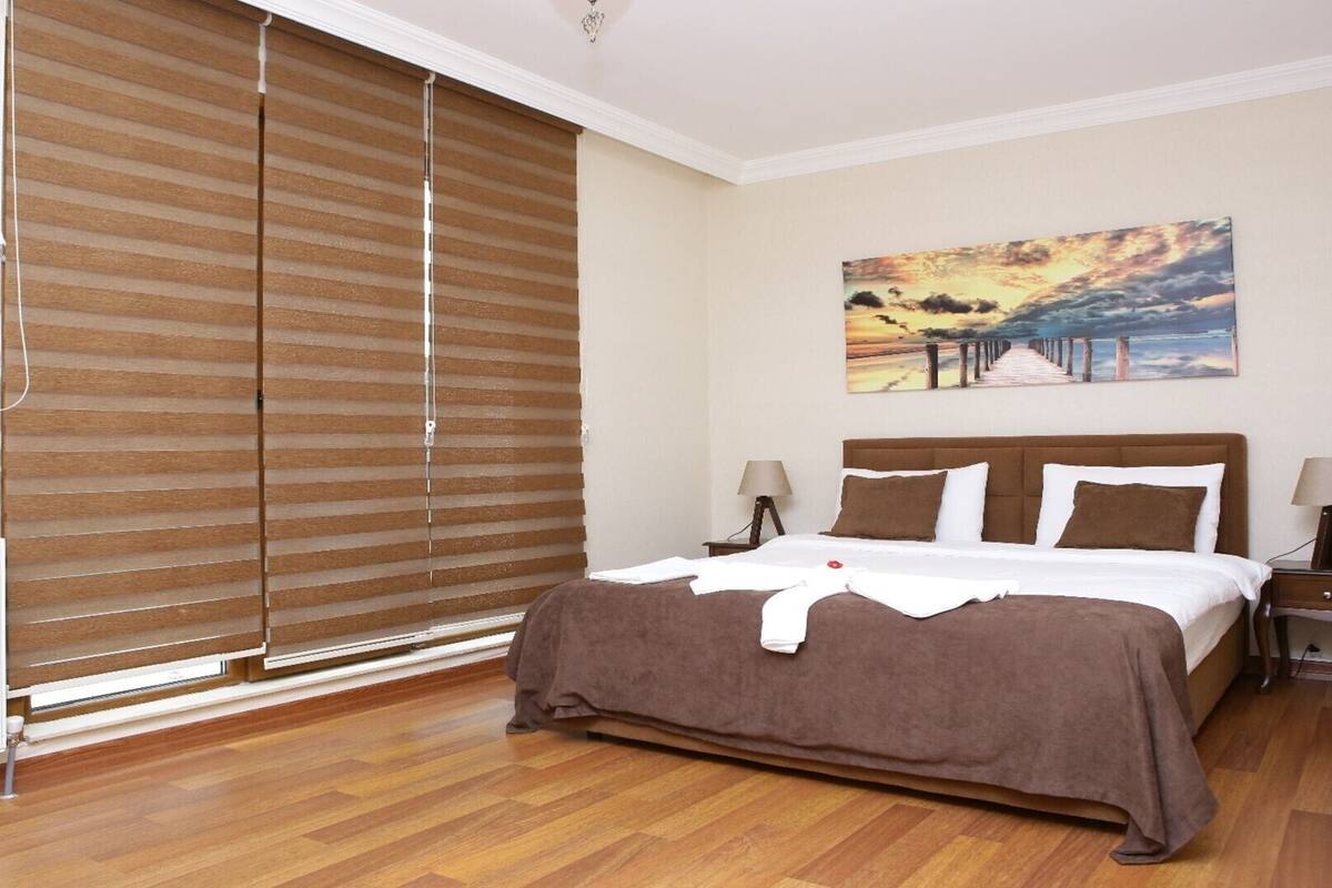 Ein Schlafzimmer mit einem großen Bett, Holzjalousien, einem Sonnenuntergangsgemälde und einem Nachttisch mit Lampe.