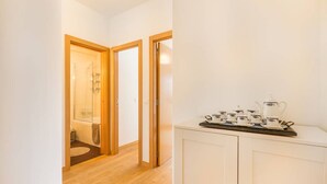 Een gang met een houten vloer die leidt naar een badkamer met een douchecabine van glas. Een wit kastje met een schaal theekopjes is zichtbaar.
