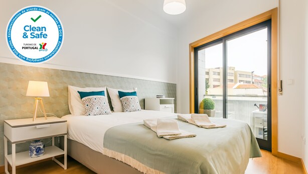 Een schone en veilige hotelkamer met een bed, nachtkastje, lamp en uitzicht op de stad door een groot raam.