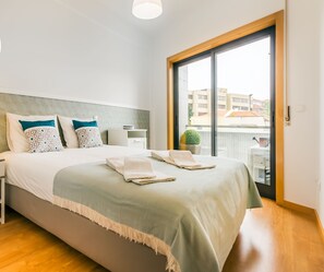 Habitació d'hotel neta i segura, amb llit, tauleta de nit, làmpada i vista de la ciutat a través d'una finestra gran.