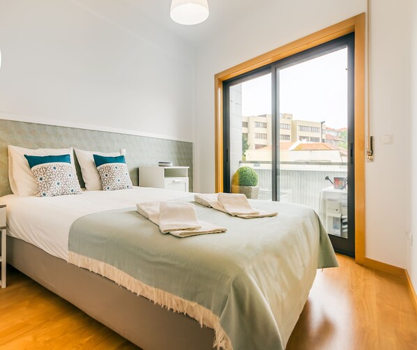 A clean and safe hotel room with a bed, nightstand, lamp, and a view of the city through a large window.