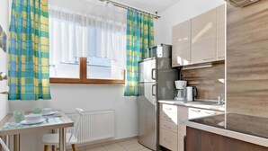 A compact kitchen with a refrigerator, microwave, and stove. There is a dining table with chairs and a window with curtains.