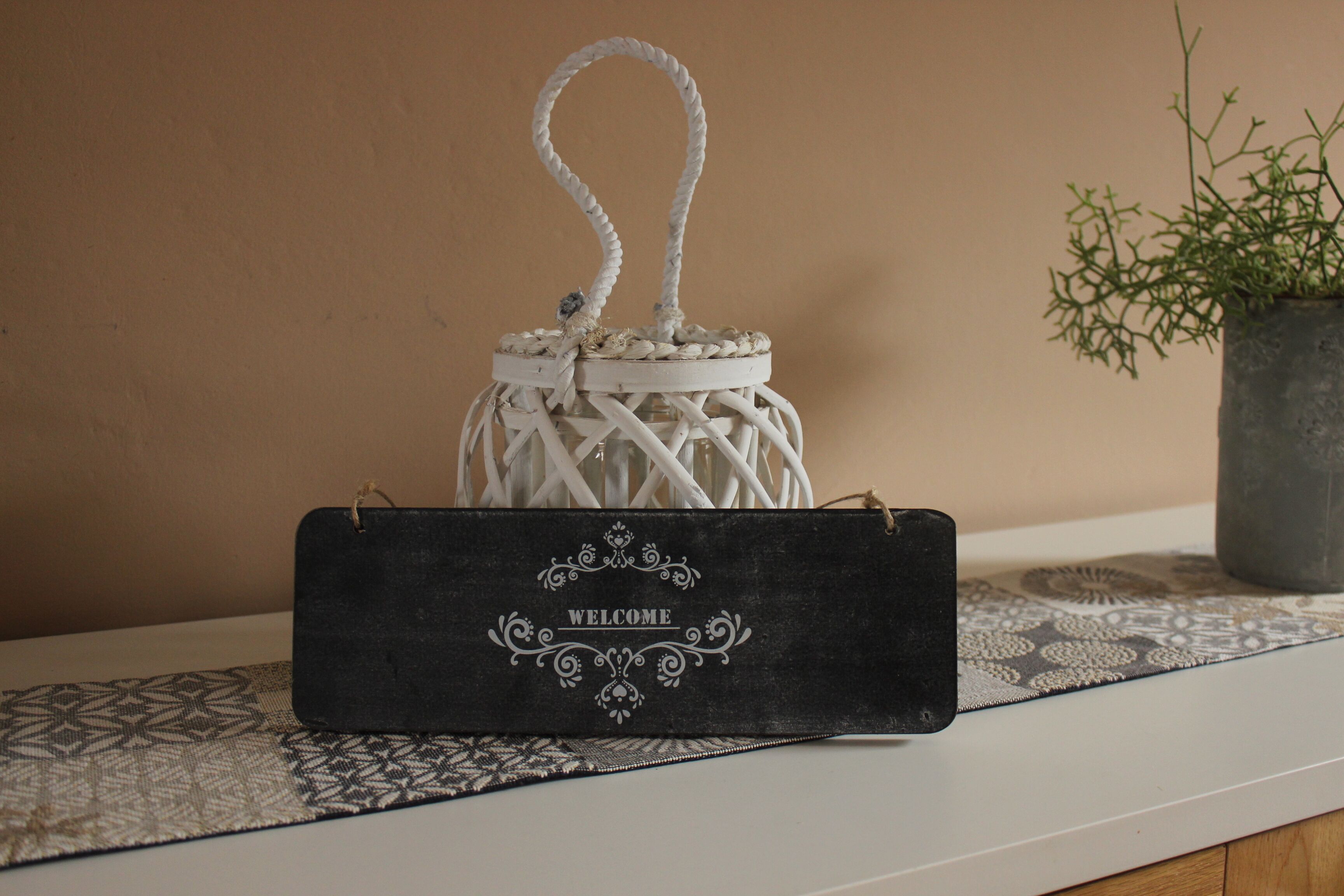 A white woven lantern with a rope handle, a black welcome sign, and a patterned table runner on a white table against a beige wall.
