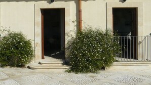 Un bâtiment doté de deux portes, d’un toit recouvert de tuiles et d’un petit jardin agrémenté de fleurs blanches.
