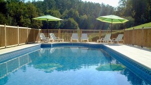 Une piscine rectangulaire dont le bord est recouvert de carrelage, entourée d’une terrasse en bois. On y trouve des chaises longues et deux parasols qui offrent de l’ombre.