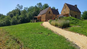 Une maison en pierre, accessible par un chemin de gravier, entourée de verdure et d’arbres.