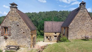 Deux bâtiments en pierre avec des toits à deux versants, un banc en bois et un chemin menant à une petite table entourée de chaises.