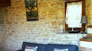Une pièce aux murs de pierre, avec un canapé bleu foncé, deux coussins, un tableau, une fenêtre avec des rideaux et un escalier.