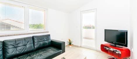 A modern living room with a black leather sofa, a wooden coffee table, a red entertainment unit with a flat-screen TV, and large windows with blinds.
