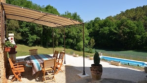 Eine Terrasse im Freien mit Tisch und Stühlen, einer Pergola und Blick auf ein Schwimmbad und bewaldete Hügel.