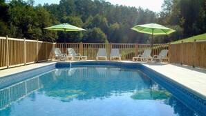 Ein rechteckiger Pool mit Fliesenrand, umgeben von einer Holzterrasse und einem Holzzaun. Bietet Liegestühle und Sonnenschirme am Pool.