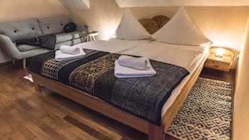 A neatly made bed with a patterned blanket, a grey sofa, and a small table with a lamp in a room with wooden flooring.