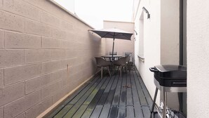 A narrow outdoor patio with a brick wall, a table with chairs, an umbrella, and a barbecue grill.