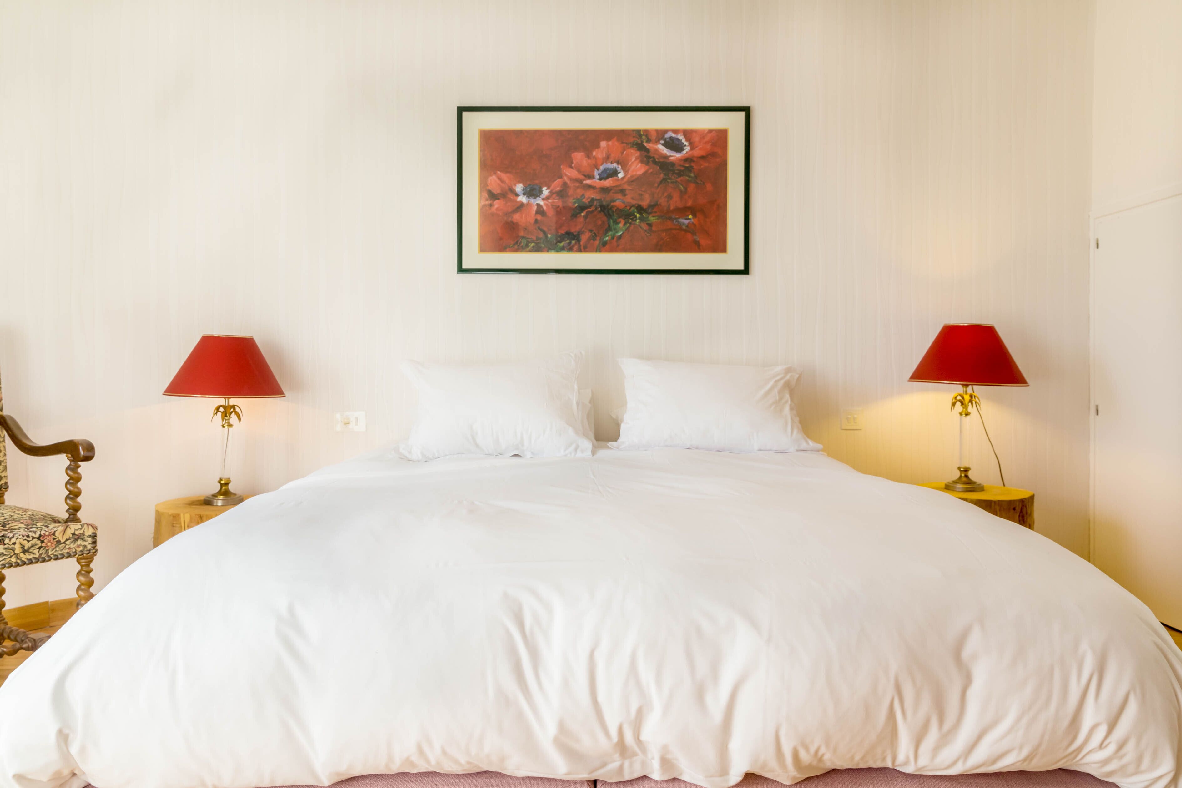 Cama bem arrumada com lençóis brancos, duas luminárias vermelhas nas mesas de cabeceira e uma obra de arte floral emoldurada acima da cama.