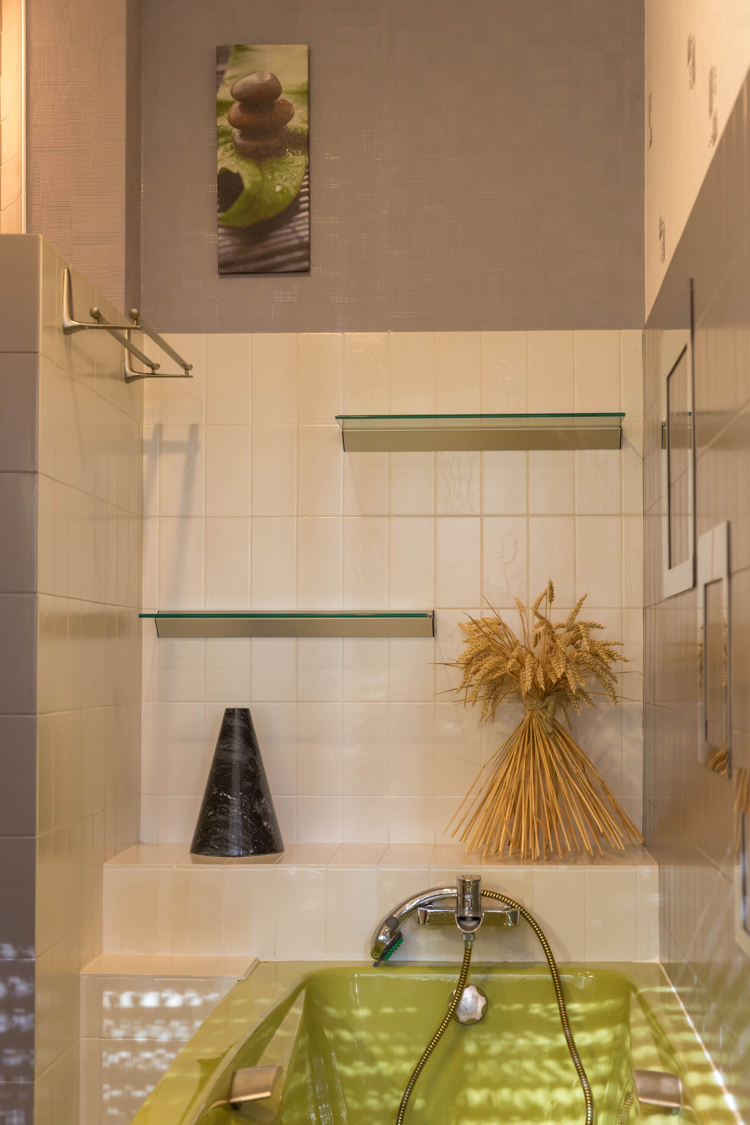 A modern bathroom with a green bathtub, a glass shelf, and a decorative vase of dried plants.