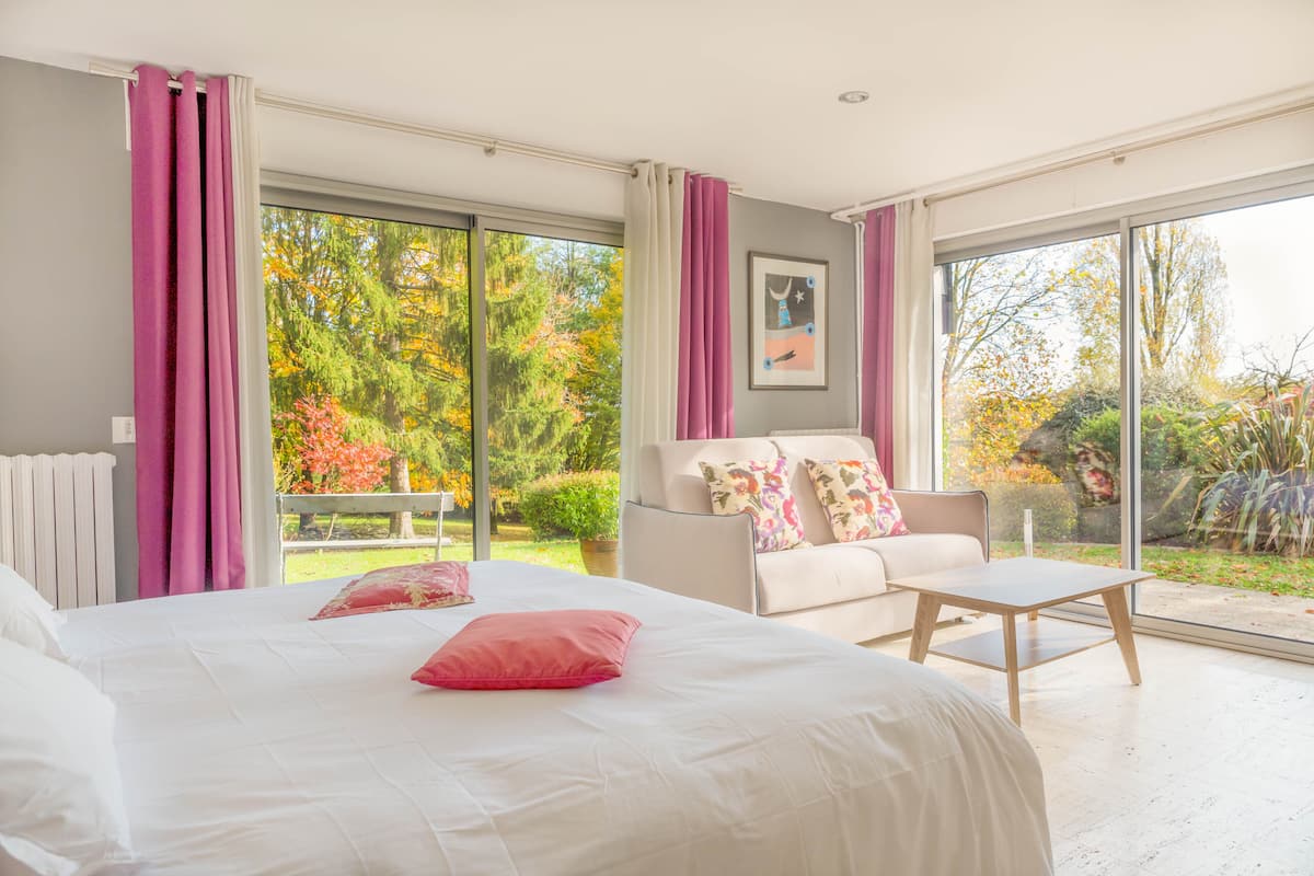 A bedroom with a large bed, a sofa, and a table. There is a view of a garden through the windows.