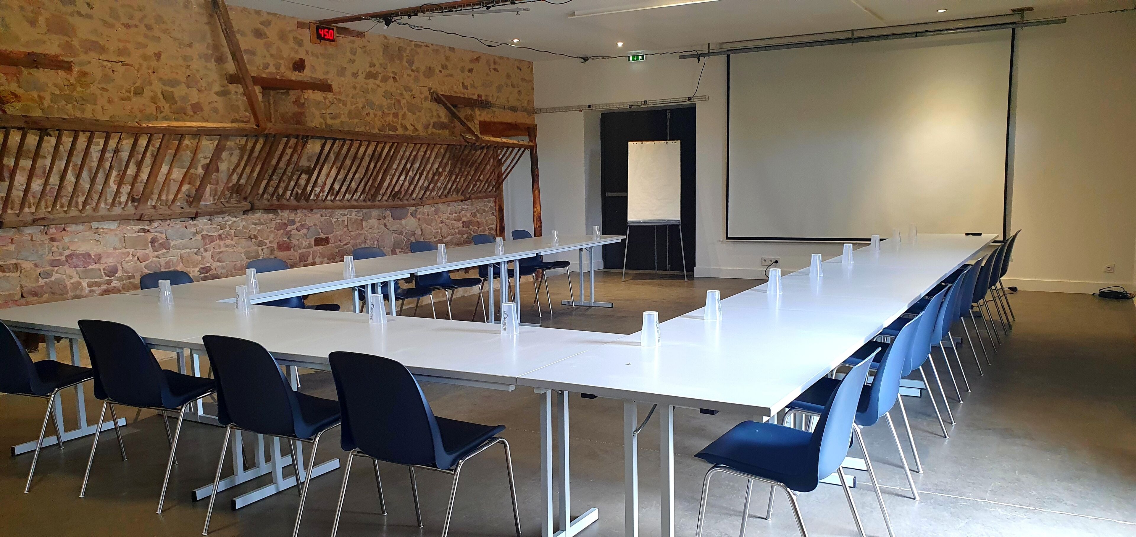 Een vergaderruimte met een lange tafel, blauwe stoelen, een whiteboard en een scherm voor een beamer.