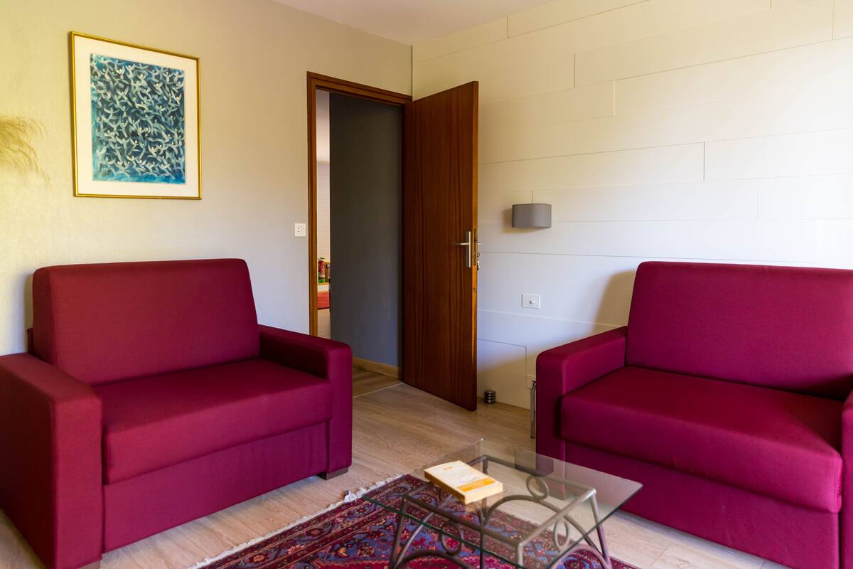 A living room with two red sofas, a glass coffee table, a patterned rug, a framed artwork on the wall, and a door leading to another room.