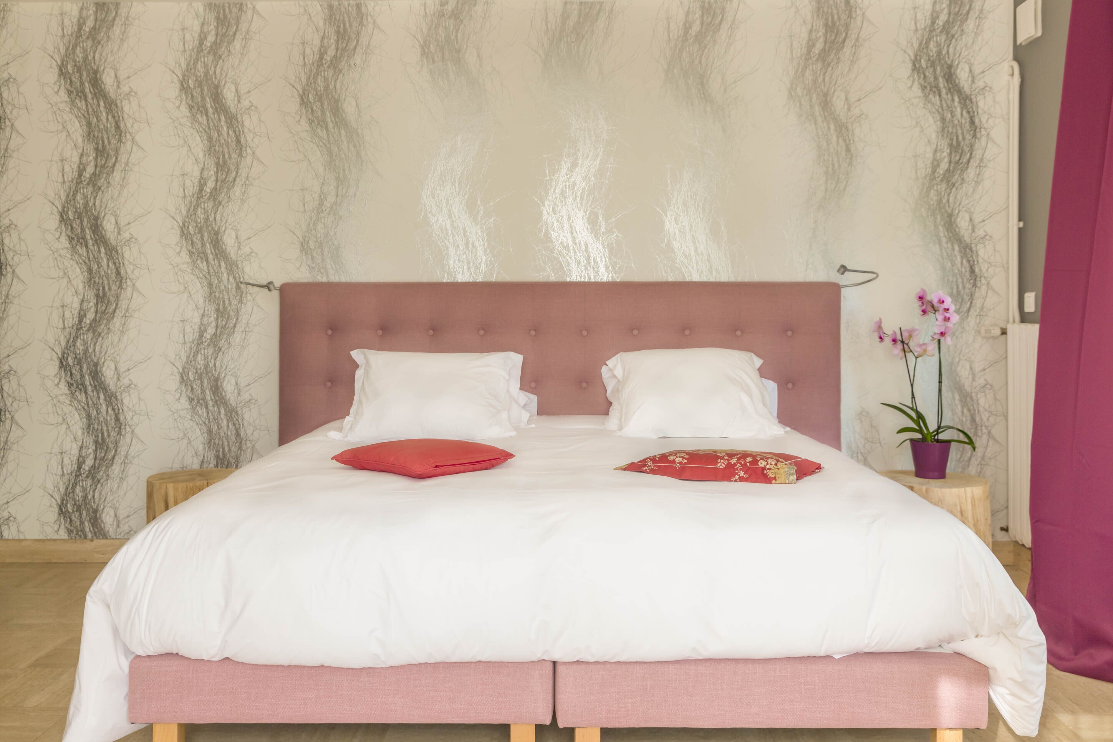 Un lit bien fait, avec une literie blanche et rouge, une tête de lit rose et une petite table de chevet en bois sur laquelle est posée une plante en pot.