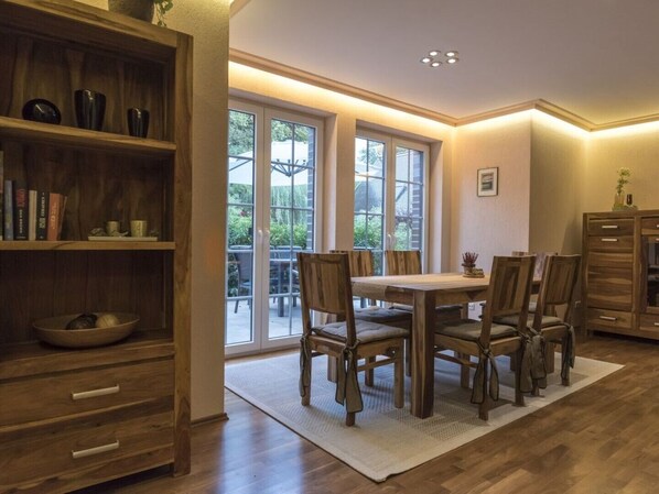 A dining area with a wooden table and chairs, a bookshelf, and a view through large windows.