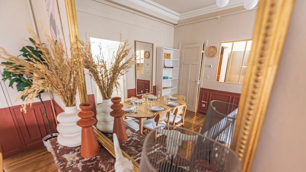 Un comedor con una mesa de madera dispuesta para cuatro, rodeada de sillas. Hay jarrones decorativos con plantas secas sobre la mesa, y un espejo grande en la pared.
