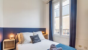 Una cama bien hecha con una colcha azul y dos toallas, una lámpara de noche y una ventana con cortinas.