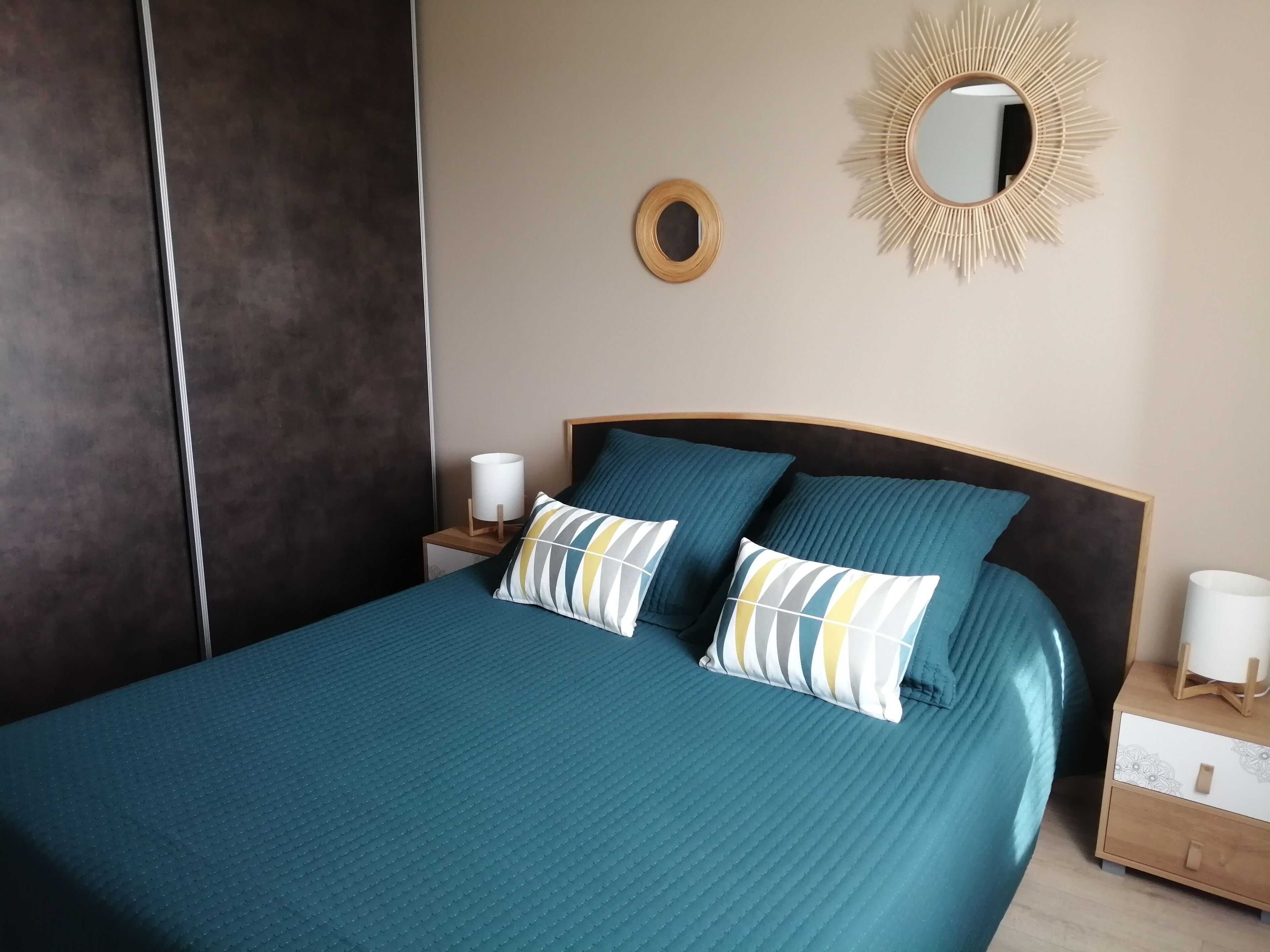 A neatly made bed with a teal bedspread and two pillows, a round mirror on the wall, and a bedside table with a lamp.
