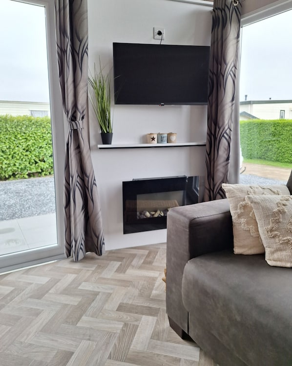 A modern living room with a grey sofa, a flat-screen TV mounted on a wall, a fireplace, and a potted plant. - NV 37 - Nieuwvliet Beach Resort (Nieuwvliet)