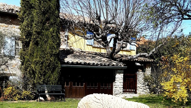 Un paysage rural avec un bâtiment en pierre, un grand arbre et des moutons qui paissent.