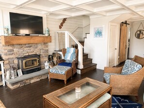 A cozy living room with a stone fireplace, wicker furniture, a flat-screen TV, and a staircase leading upstairs.