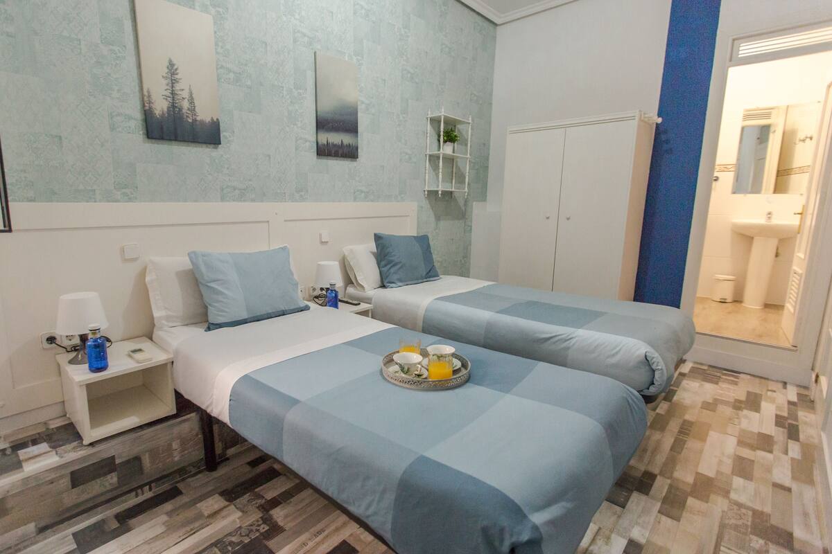 A hotel room with two beds, a nightstand, a small table with a tray of breakfast, a lamp, and a shelf with a plant.