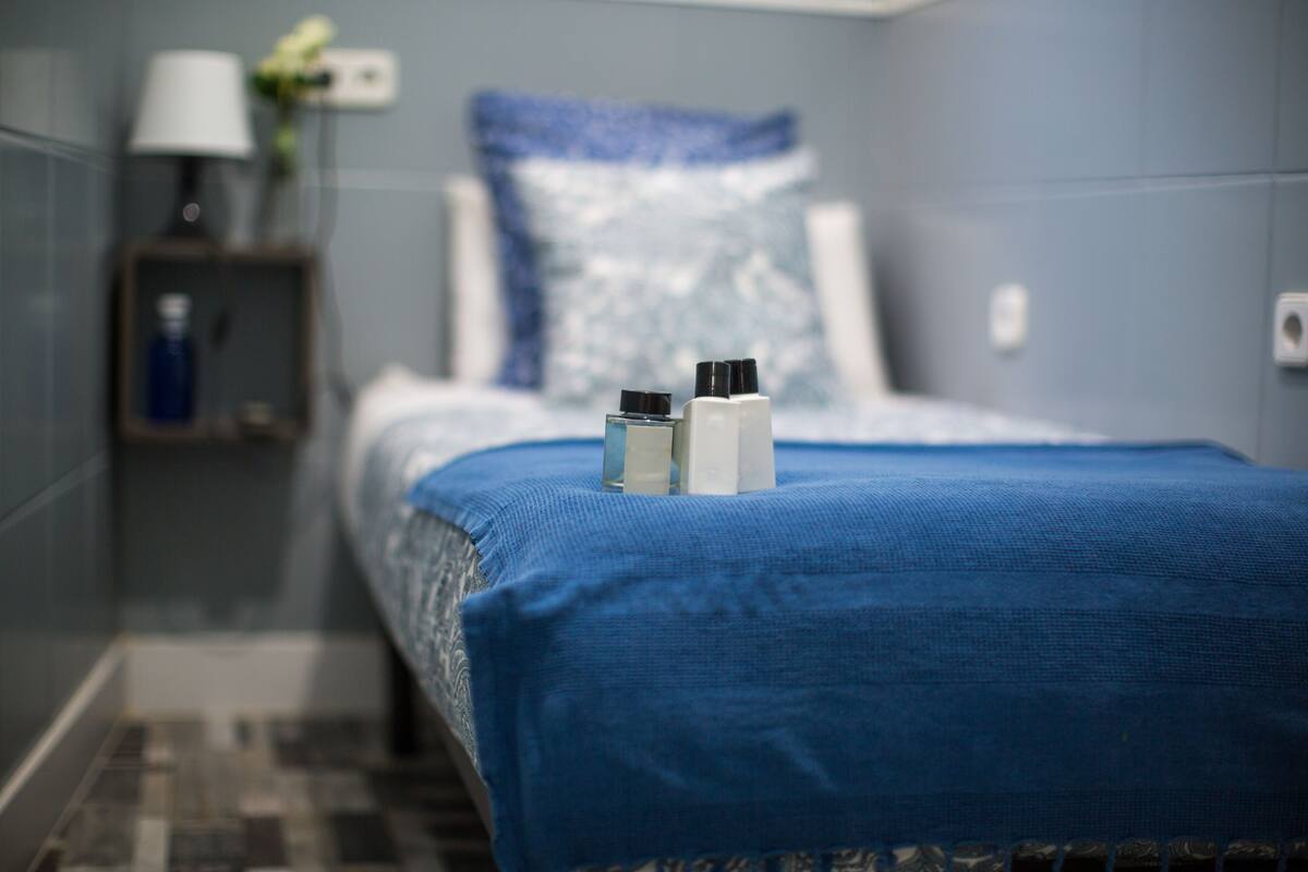 A hotel room with a neatly made bed, a nightstand with a lamp and a vase, and a blue bedspread.
