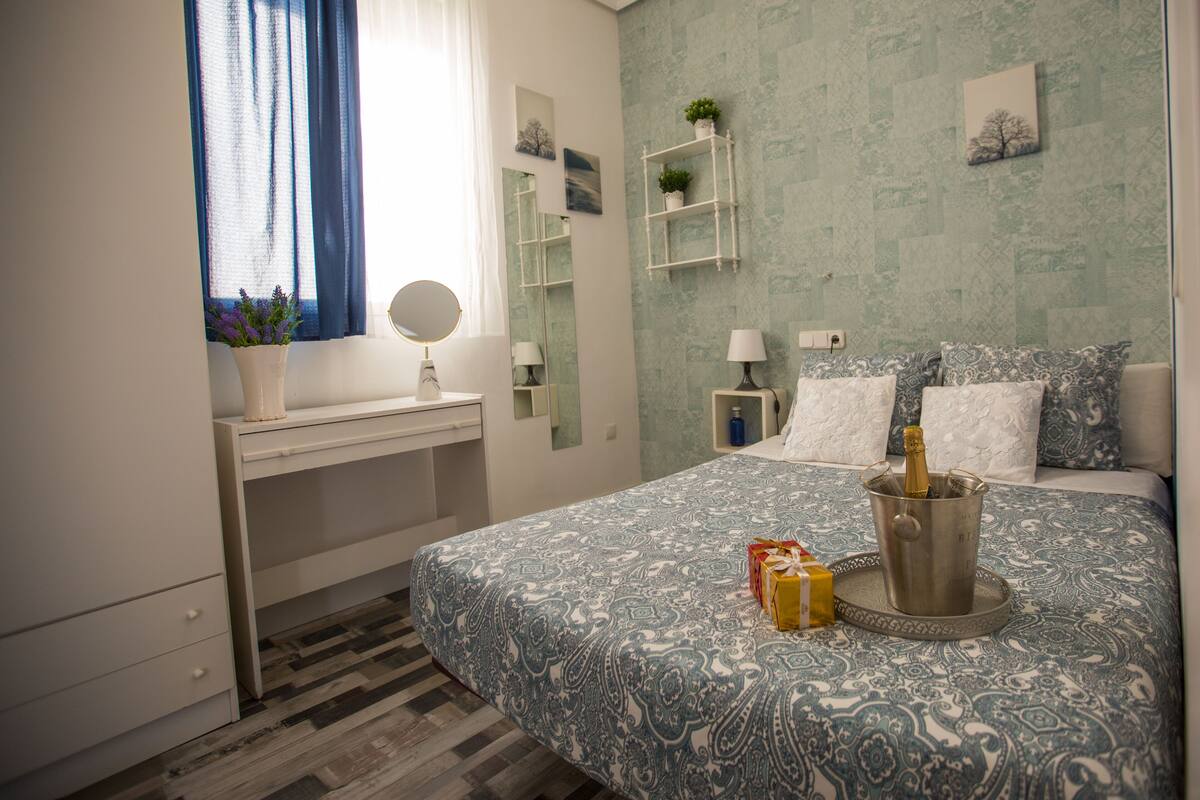 A bedroom with a bed, a desk, a chair, a window with curtains, a bedside table, a nightstand, a lamp, a mirror, a wall-mounted shelf with plants, and a bedside table with a bottle and a wrapped gift.