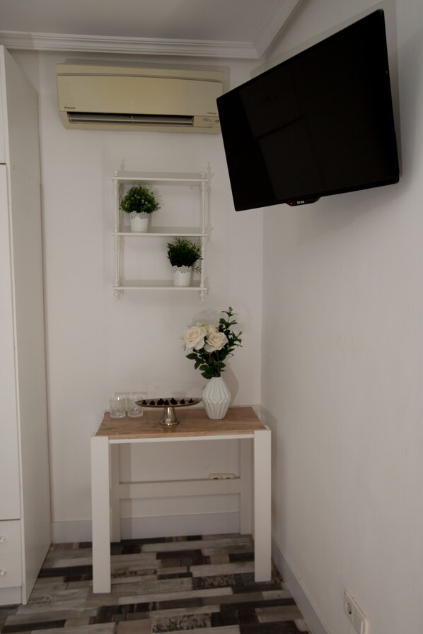 Quarto com mesa de console branca, televisão de tela plana na parede, ar-condicionado fixado na parede e prateleira com plantas em vasos.