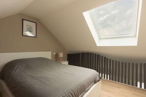 A bedroom with a sloped ceiling, a bed, a bedside table with a lamp, a framed picture on the wall, and a window with a view of the sky.