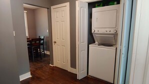 Une buanderie équipée d’un lave-linge et d’un sèche-linge, une porte donnant sur un coin repas et un couloir avec un plancher en bois.