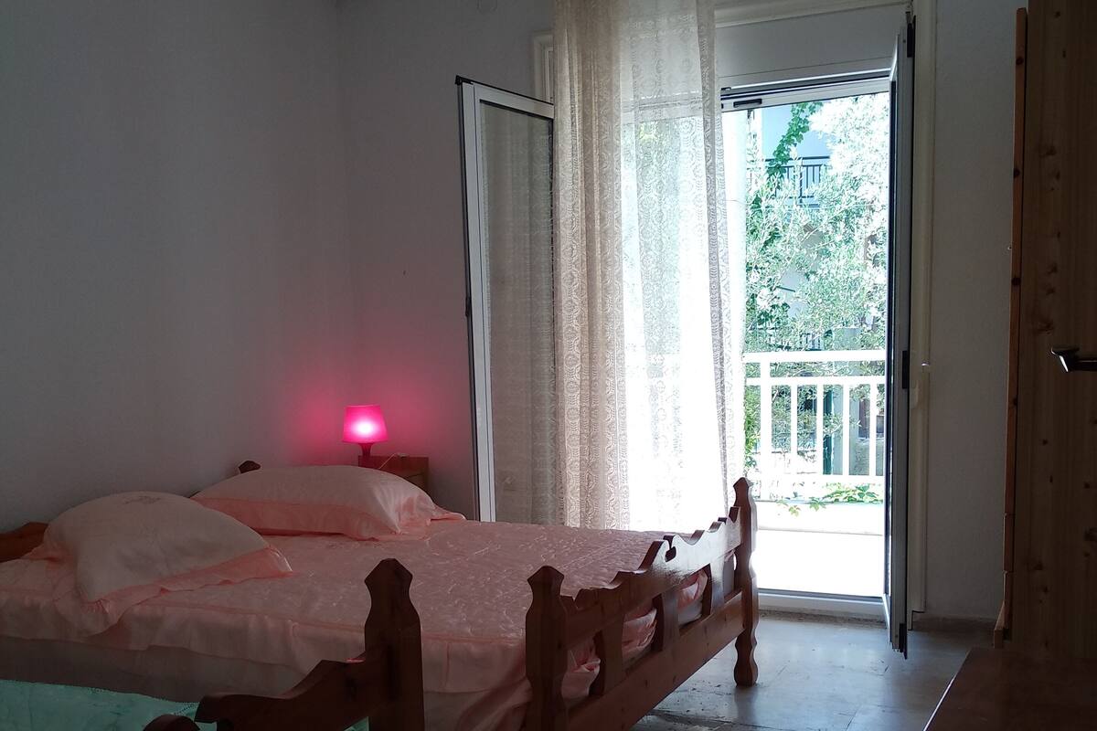A bedroom with a wooden bed, a bedside lamp, an air conditioner, and a view of the outdoors through a door.