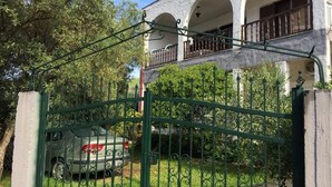 Una entrada con portón, un coche estacionado en el interior, un edificio con ventanas y balcones de arco, y una valla verde.