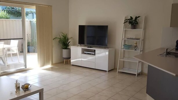Uma sala de estar moderna com televisão de tela plana, um rack branco, uma mesa de jantar e uma porta de vidro deslizante que dá para um pátio.