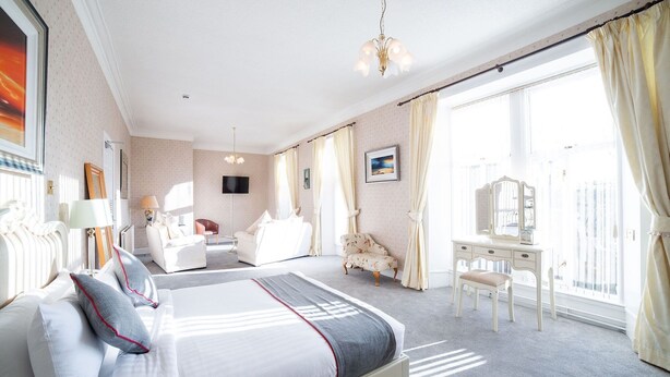 A hotel room with a large bed, a sitting area with a chair and a small table, a vanity with a mirror, and a balcony with curtains. - The Douglas Hotel (Kilmarnock)